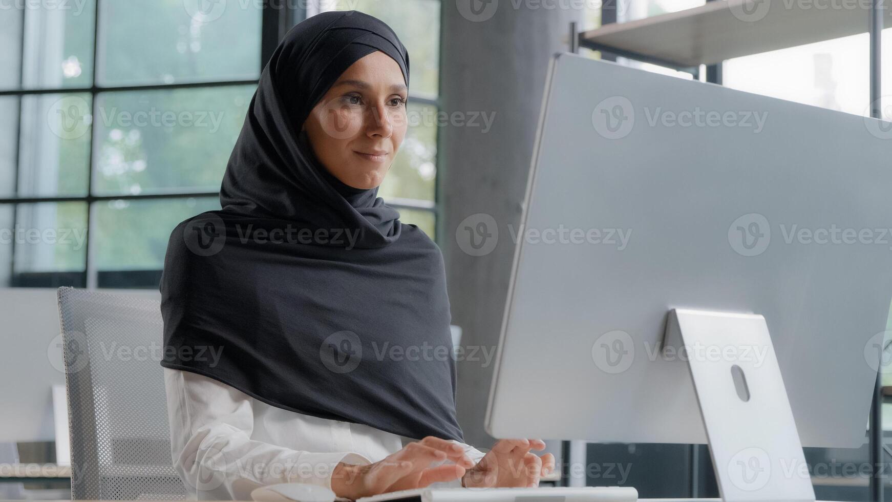 Young arab businesswoman in hijab working on computer smiling enjoying office work successful woman professional manager company employee develops project online searching for information on Internet photo