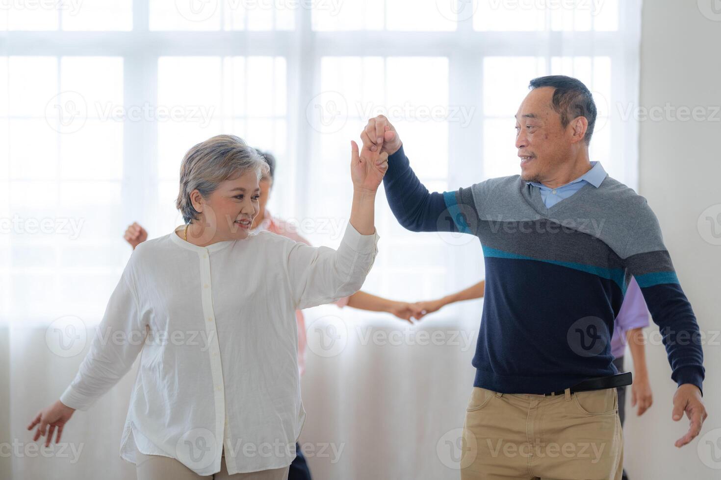 Asian Older male and females people dance with their partners on a dancing floor in living space. Happy older couple performing get exercise. Joyful carefree retired senior friends enjoying relaxation photo