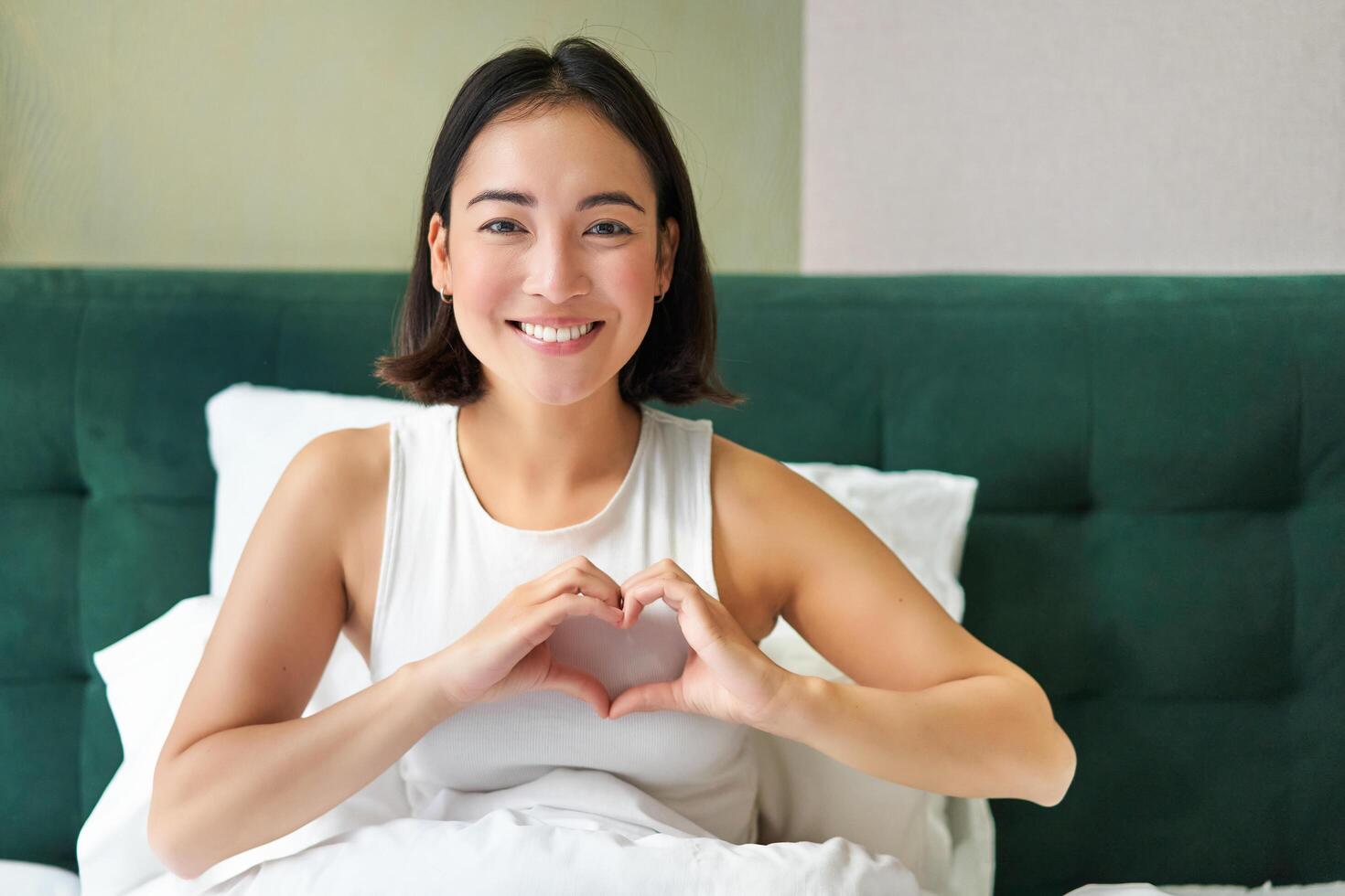 sonriente coreano niña muestra corazón, amor firmar, gasto hora en dormitorio, acostado en cama en mañana, cubierto con blanco edredones foto