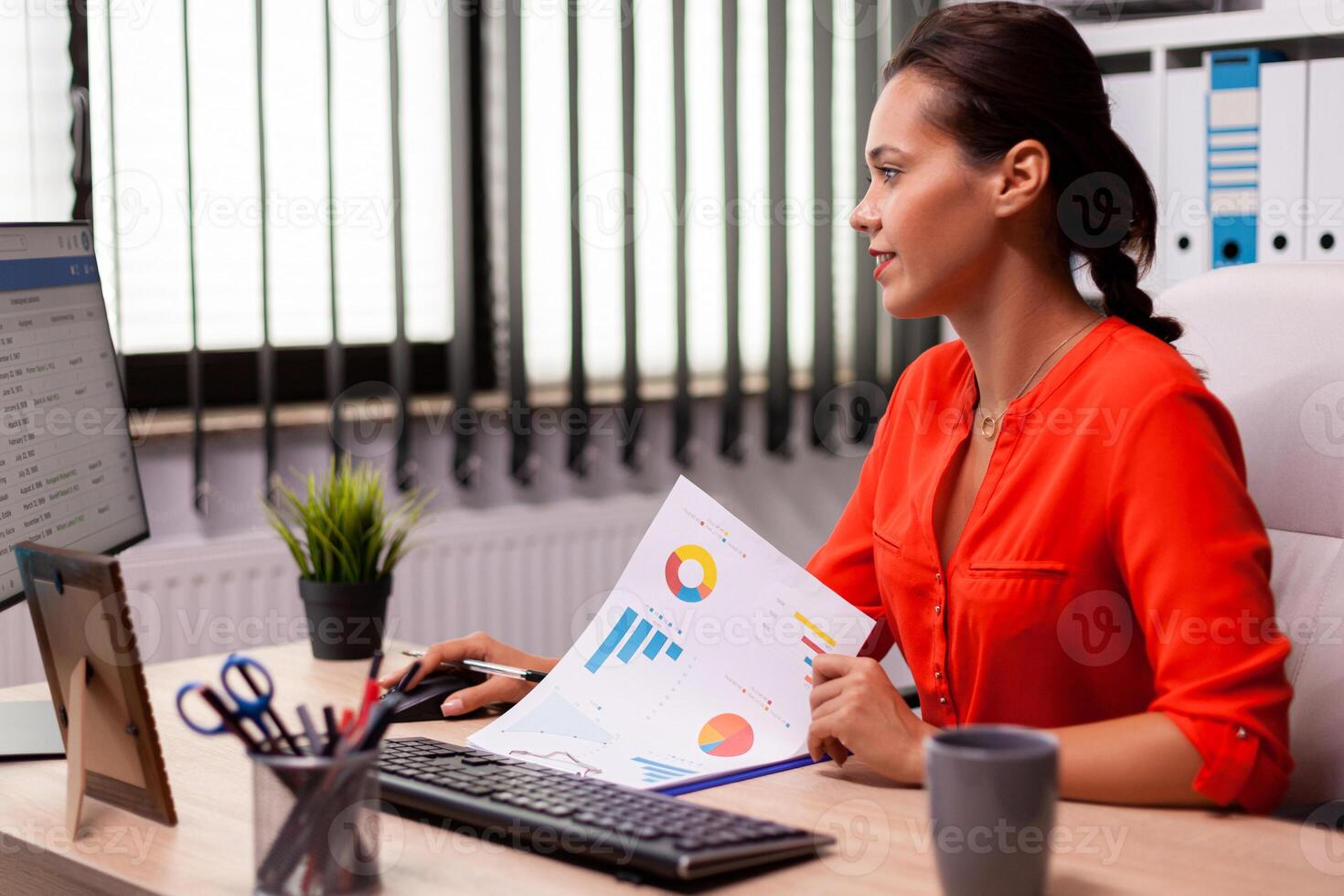 Businesswoman executive leadership planing financial analysis working at computer sitting at desk wearing red. Successful financial auditor in data marteking looking at graphs on computer screen and writing report. photo