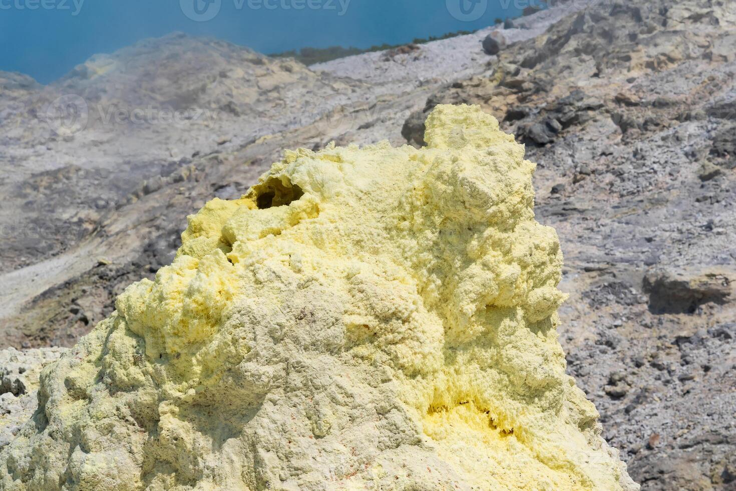 cone of sulfur deposits around a fumarole in a solfataric field illuminated by the sun against a cloudy landscape in the distance photo