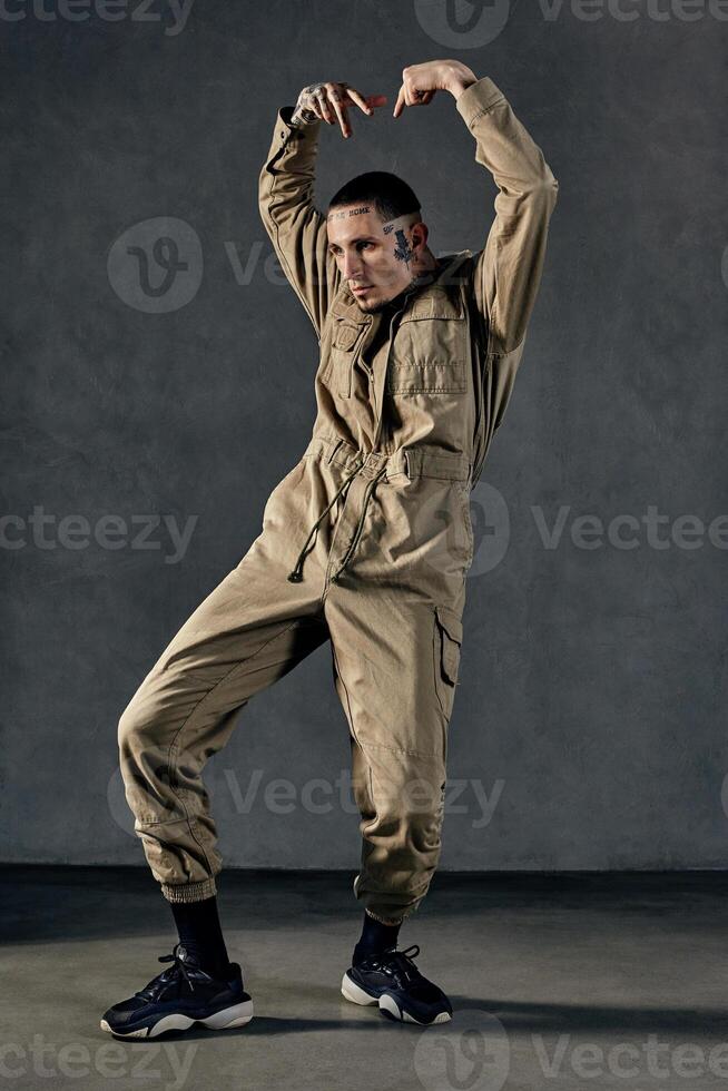 Active guy with tattooed body and face, earrings, beard. Dressed in khaki overalls and black sneakers. Dancing on gray background. Dancehall, hip-hop photo