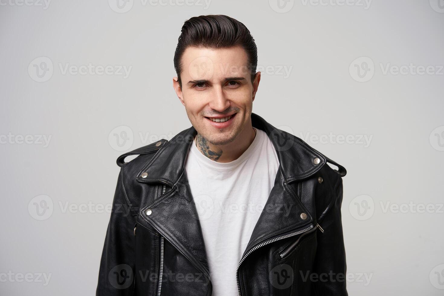 Flirting, provocative, definitely, attractively smiling rocker man, rebel, motto racer, stylish hairstyle in briolin, looks in the style of 50s-60s in black leather jacket, over white background photo