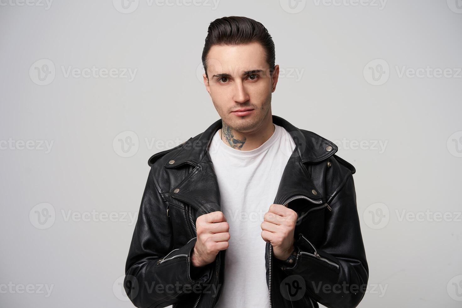 Close up of attractive rebel, brutal with a stylish haircut and styling with a grease, tattoo on the neck,keeps hands like putting on black rocker-style leather jacket, over white background photo