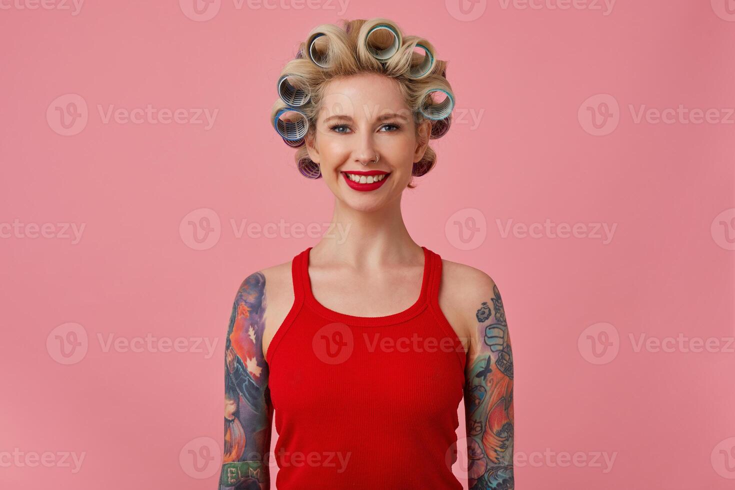 Cheerful young blonde tattooed female with evening makeup making hairdo while posing over pink background with hands down, looking at camera with charming smile photo