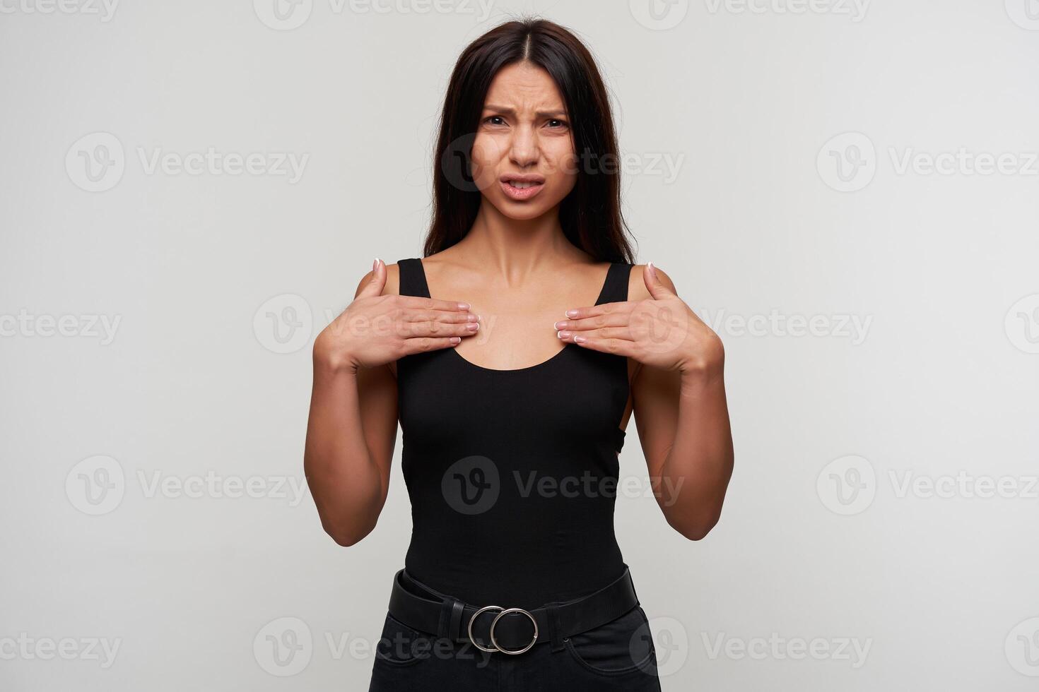 Displeased young long haired brunette female holding palms on her chest and looking at camera with pout, frowning her face while posing over white background photo