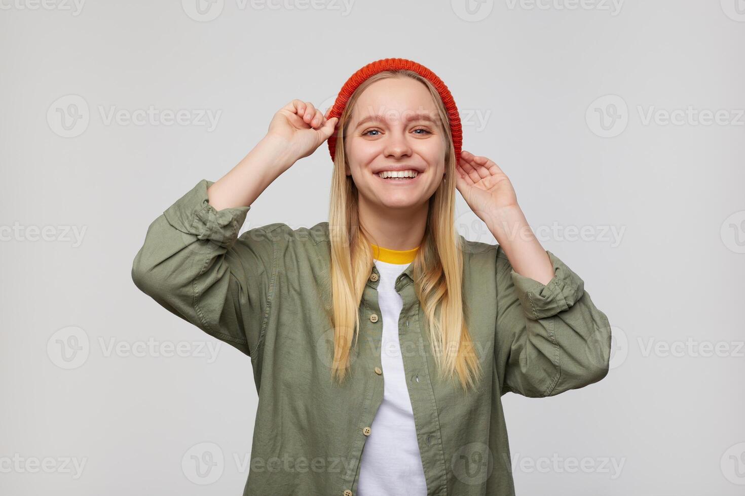 Cheerful young lovely long haired white-headed female keeping raised hands on her red hat while looking joyfully at camera with broad smile, isolated over blue background photo