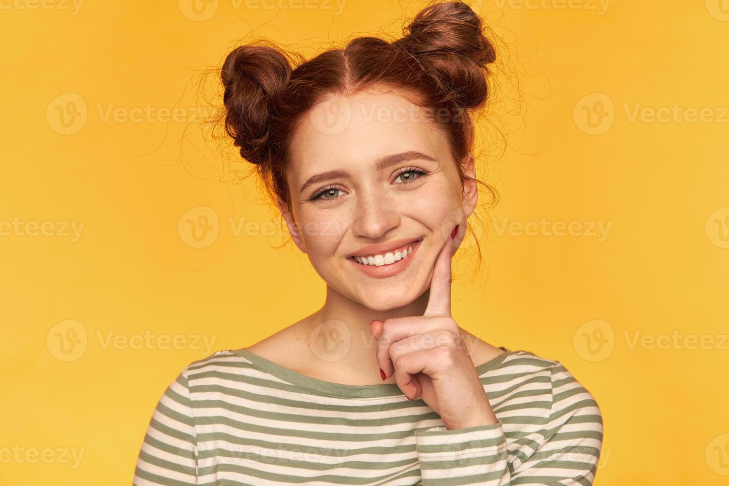 Portrait of attractive, red hair girl with two buns. Looking playful and touching her cheek. Wearing striped sweater and watching at the camera isolated, closeup over yellow background photo