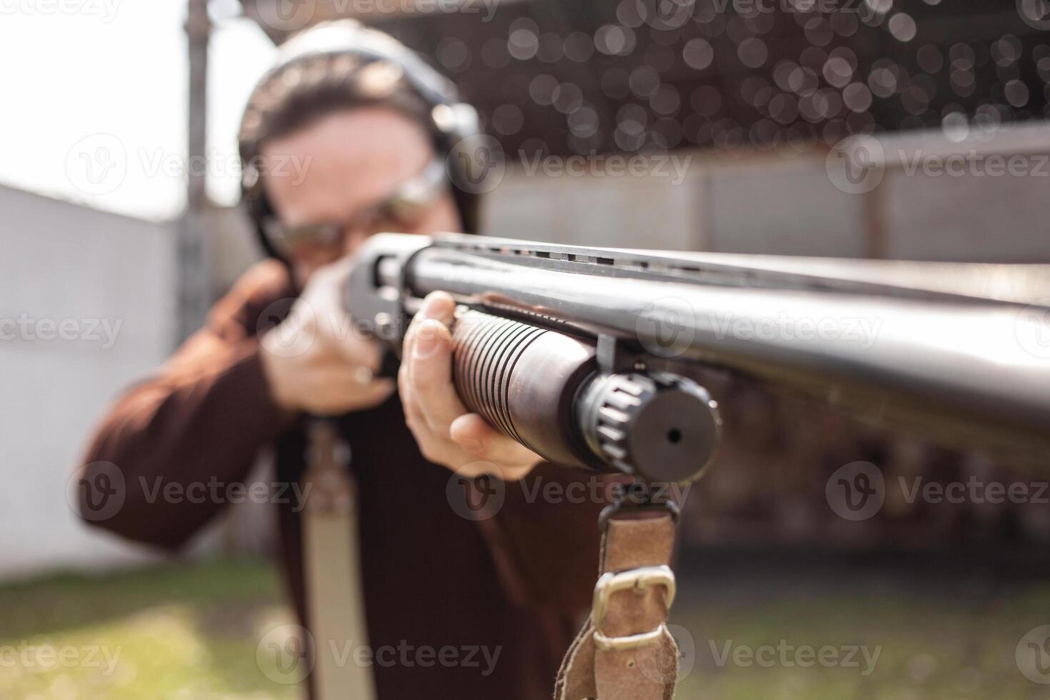A man in protective glasses and headphones. A pump-action shotgun. Tyre's outdoors. A wall and a roof with bullet holes. Sport photo