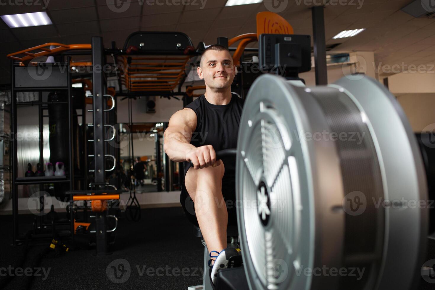 handsome male coach sits on a simulator in gym. Imitation rowing, boat with oars, for rowers. Smiling, happy, sports active healthy lifestyle, indoor concept, coaching and individual lessons. portrait photo
