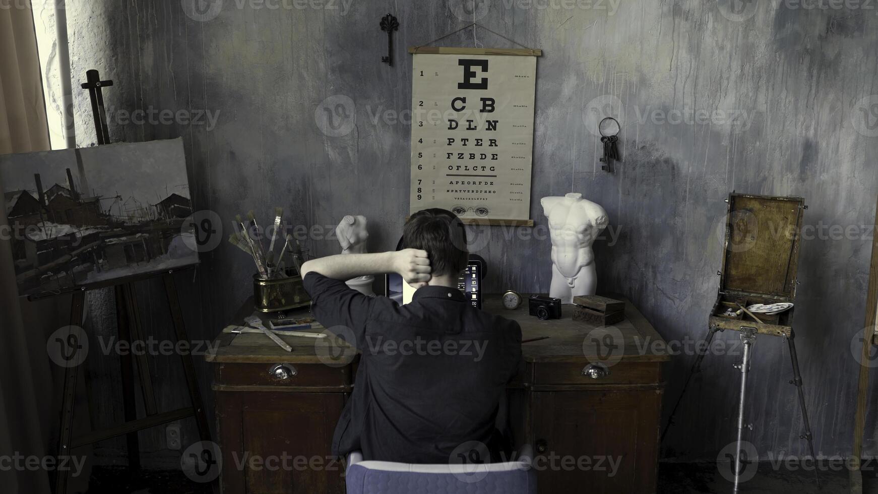 Rear view of a man working on computer in the art studio with a painting, plaster sculptures, and brushes on concrete stylized wall background. Stock footage. Male in the art workshop. photo