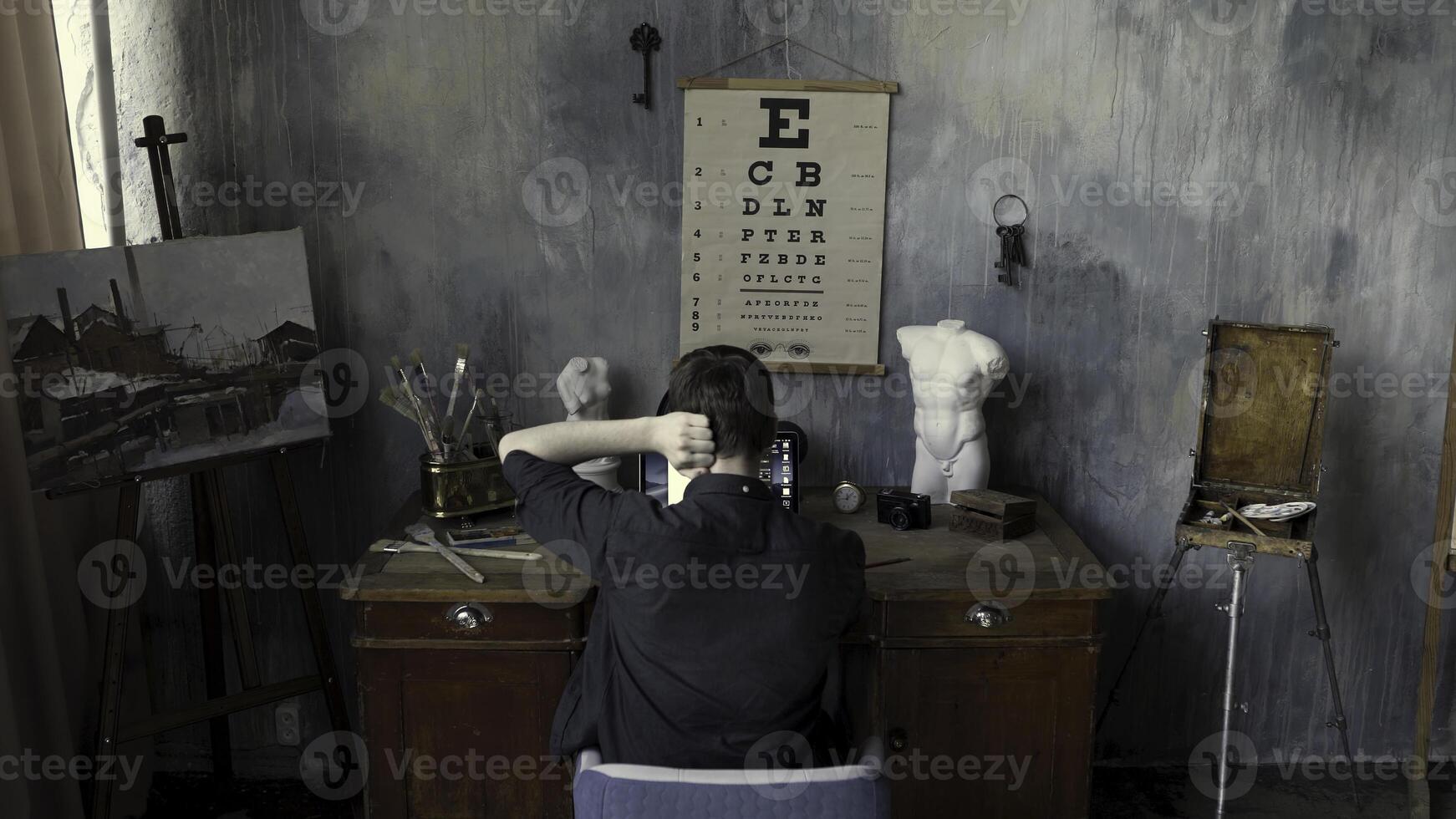 posterior ver de un hombre trabajando en computadora en el Arte estudio con un cuadro, yeso esculturas, y cepillos en hormigón estilizado pared antecedentes. valores imágenes. masculino en el Arte taller. foto