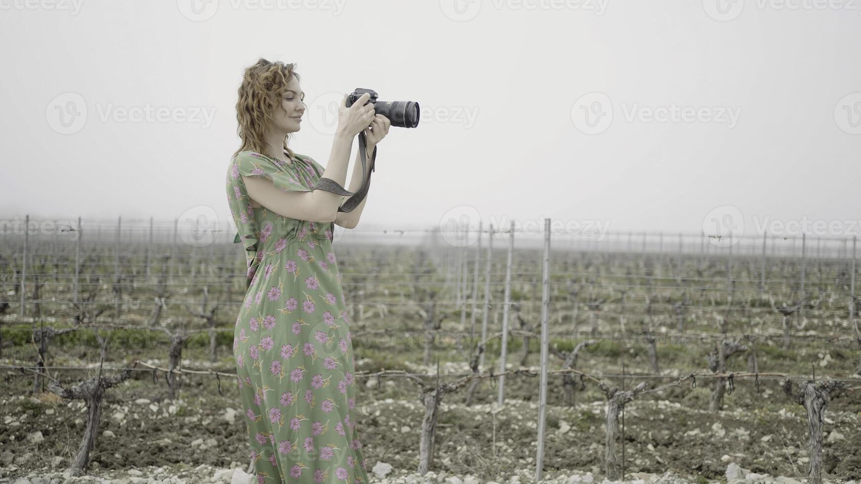 Young pretty woman takes pictures in harvest field. Action. Side view of female photographer in a field on grey sky background. photo