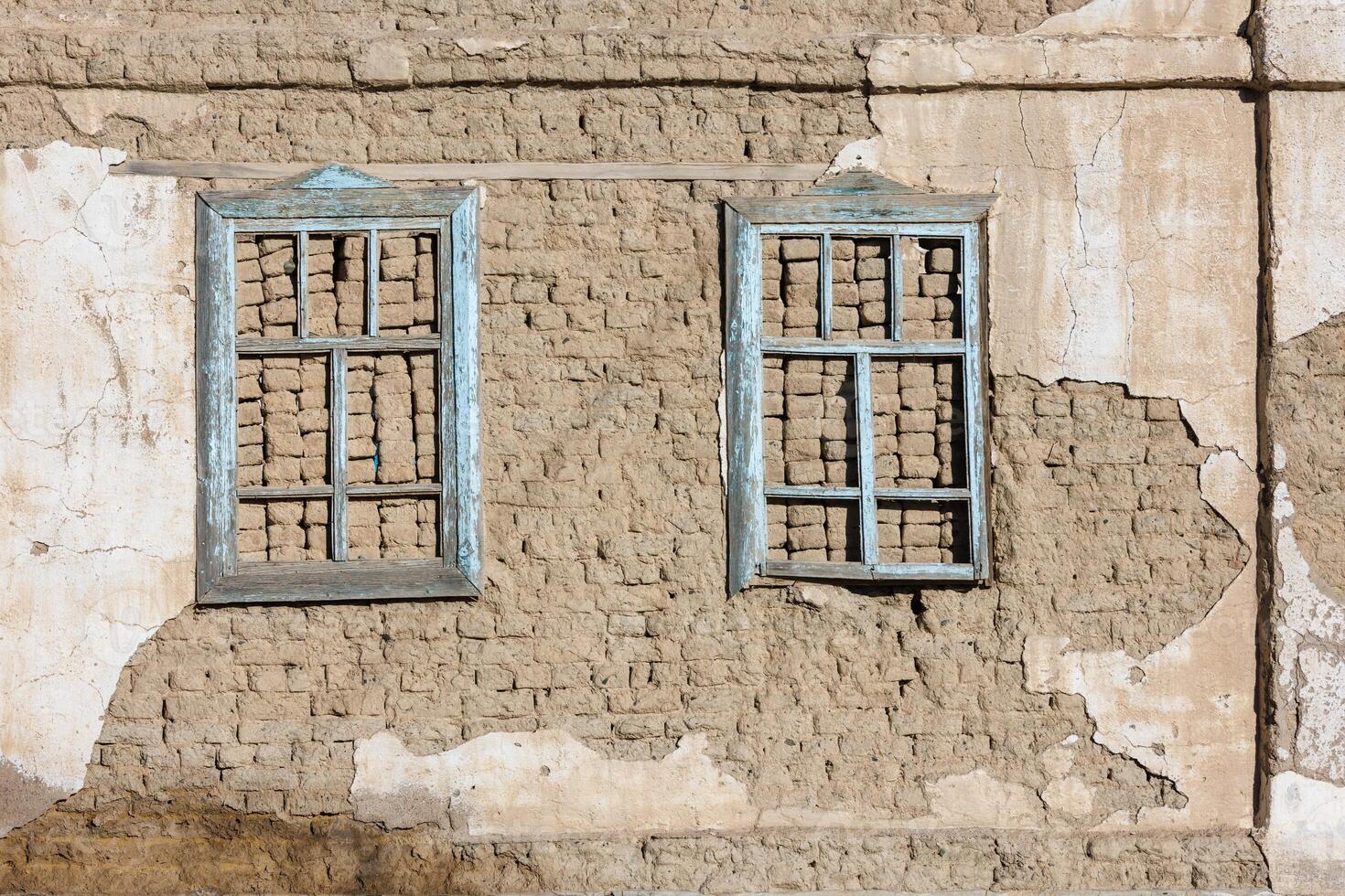 old house adobe air brick wall with two embedded wooden window frames and peeled off plaster leftovers - flat texture and full frame background photo