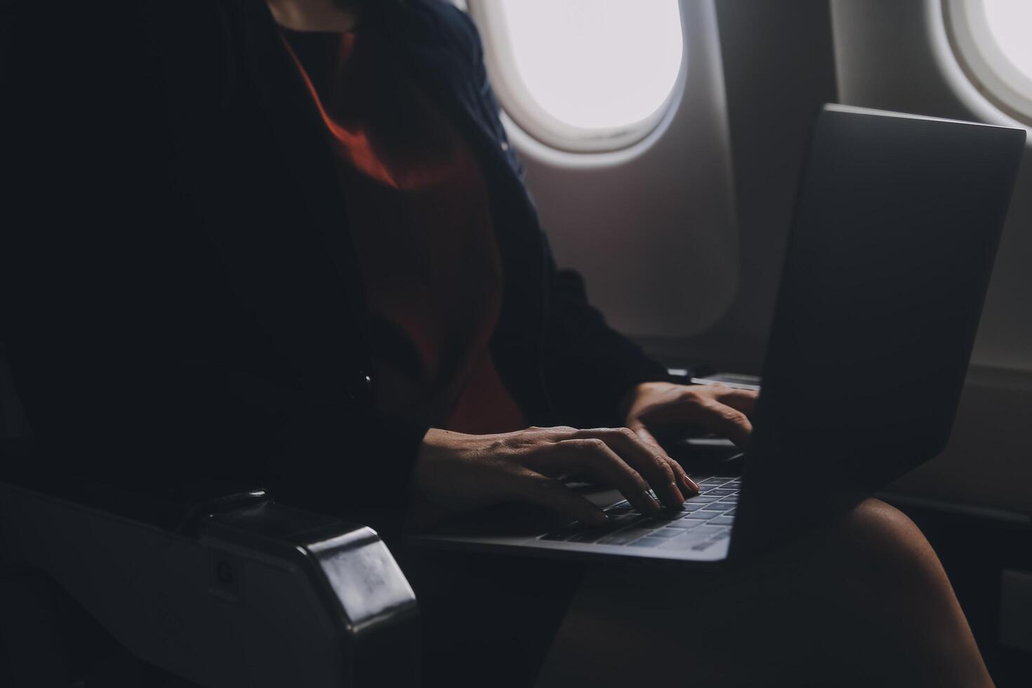 Blonde female tourist checking incoming notification on smartphone sitting on seat of airplane with netbook.Young businesswoman share media from telephone on laptop computer during plane flight photo
