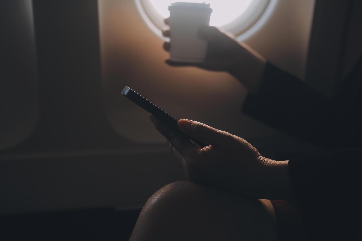 Blonde female tourist checking incoming notification on smartphone sitting on seat of airplane with netbook.Young businesswoman share media from telephone on laptop computer during plane flight photo