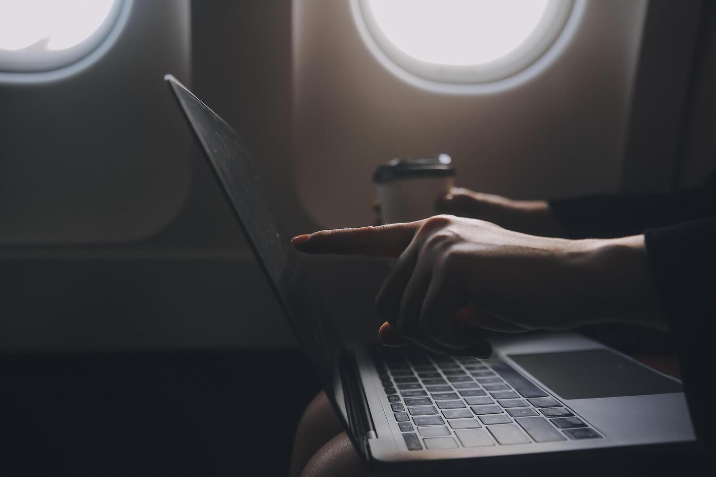 Blonde female tourist checking incoming notification on smartphone sitting on seat of airplane with netbook.Young businesswoman share media from telephone on laptop computer during plane flight photo