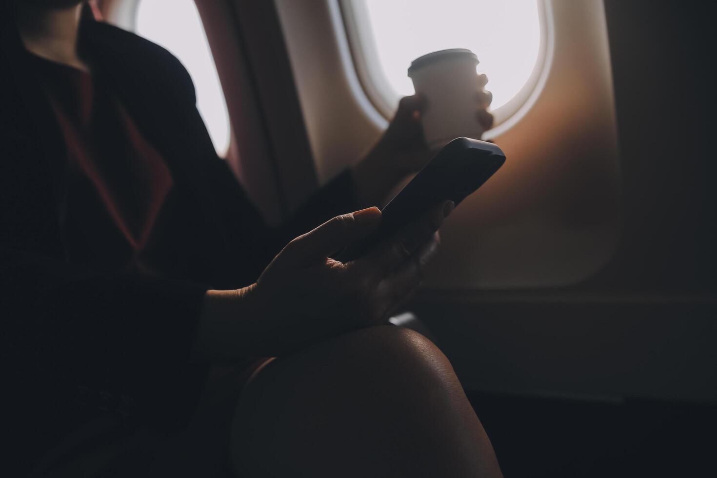 Blonde female tourist checking incoming notification on smartphone sitting on seat of airplane with netbook.Young businesswoman share media from telephone on laptop computer during plane flight photo