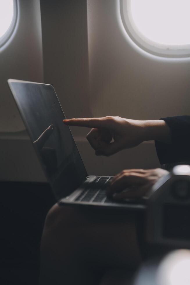 Blonde female tourist checking incoming notification on smartphone sitting on seat of airplane with netbook.Young businesswoman share media from telephone on laptop computer during plane flight photo