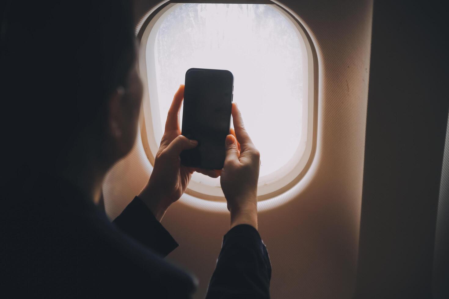 Blonde female tourist checking incoming notification on smartphone sitting on seat of airplane with netbook.Young businesswoman share media from telephone on laptop computer during plane flight photo