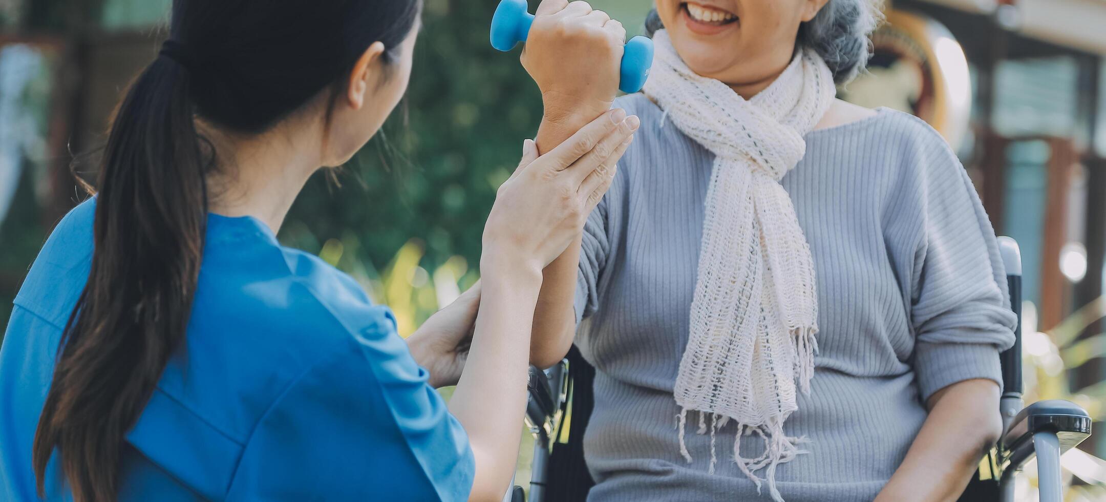 Young nurse or physiotherapist in scrubs helping a happy retired old woman do fitness exercises with light weight dumbbells at home. Concept of physiotherapy for seniors photo