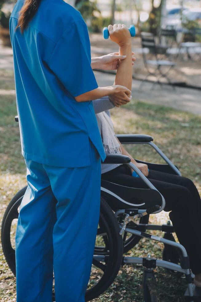 Young nurse or physiotherapist in scrubs helping a happy retired old woman do fitness exercises with light weight dumbbells at home. Concept of physiotherapy for seniors photo