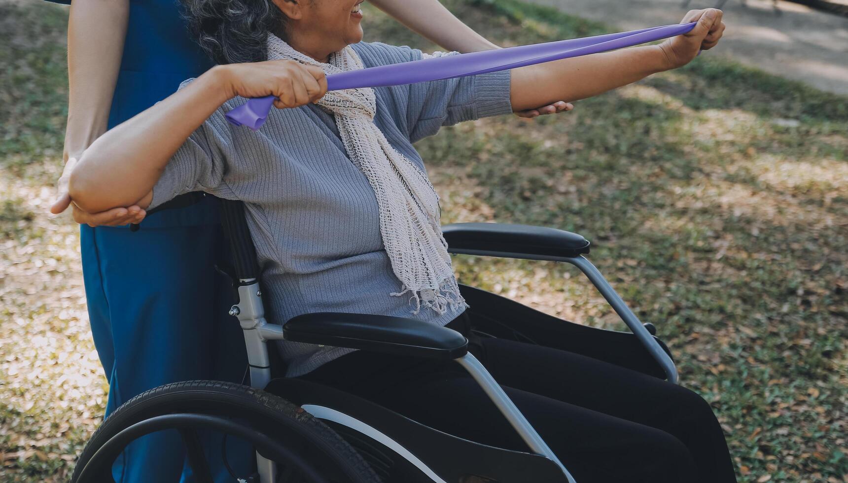 Young nurse or physiotherapist in scrubs helping a happy retired old woman do fitness exercises with light weight dumbbells at home. Concept of physiotherapy for seniors photo