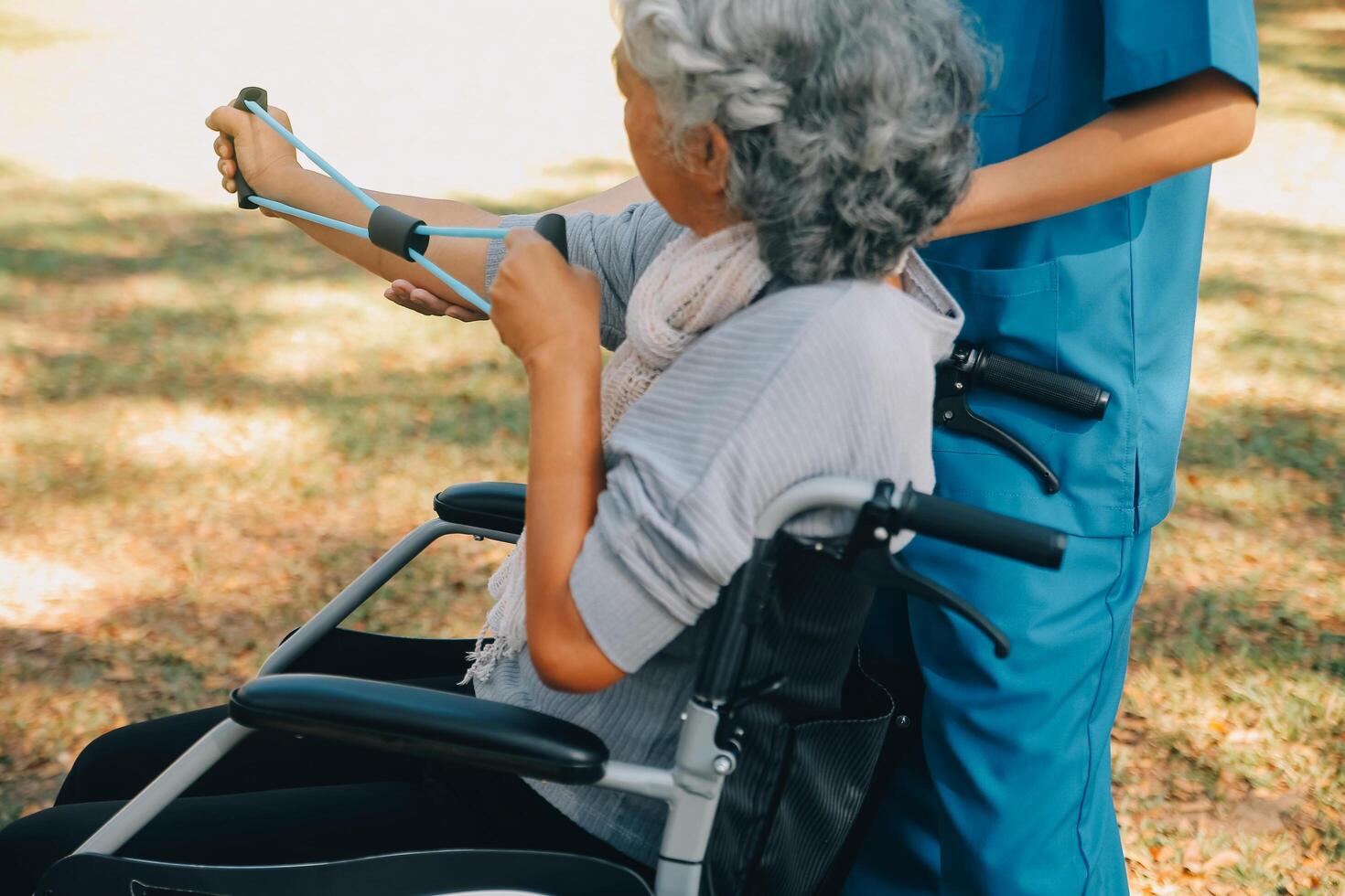 Young nurse or physiotherapist in scrubs helping a happy retired old woman do fitness exercises with light weight dumbbells at home. Concept of physiotherapy for seniors photo