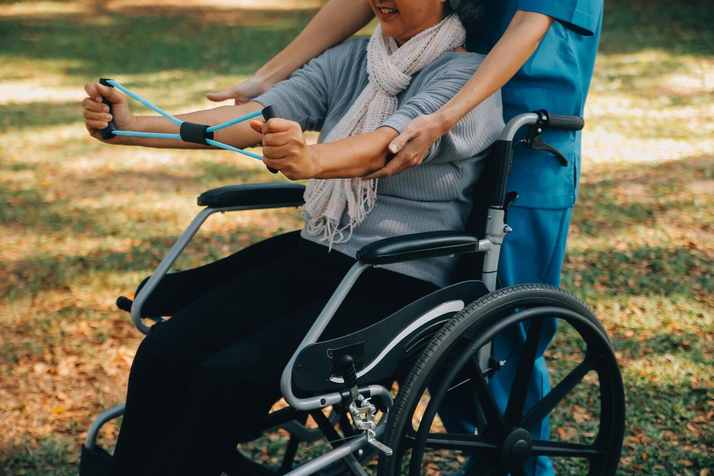 Young nurse or physiotherapist in scrubs helping a happy retired old woman do fitness exercises with light weight dumbbells at home. Concept of physiotherapy for seniors photo