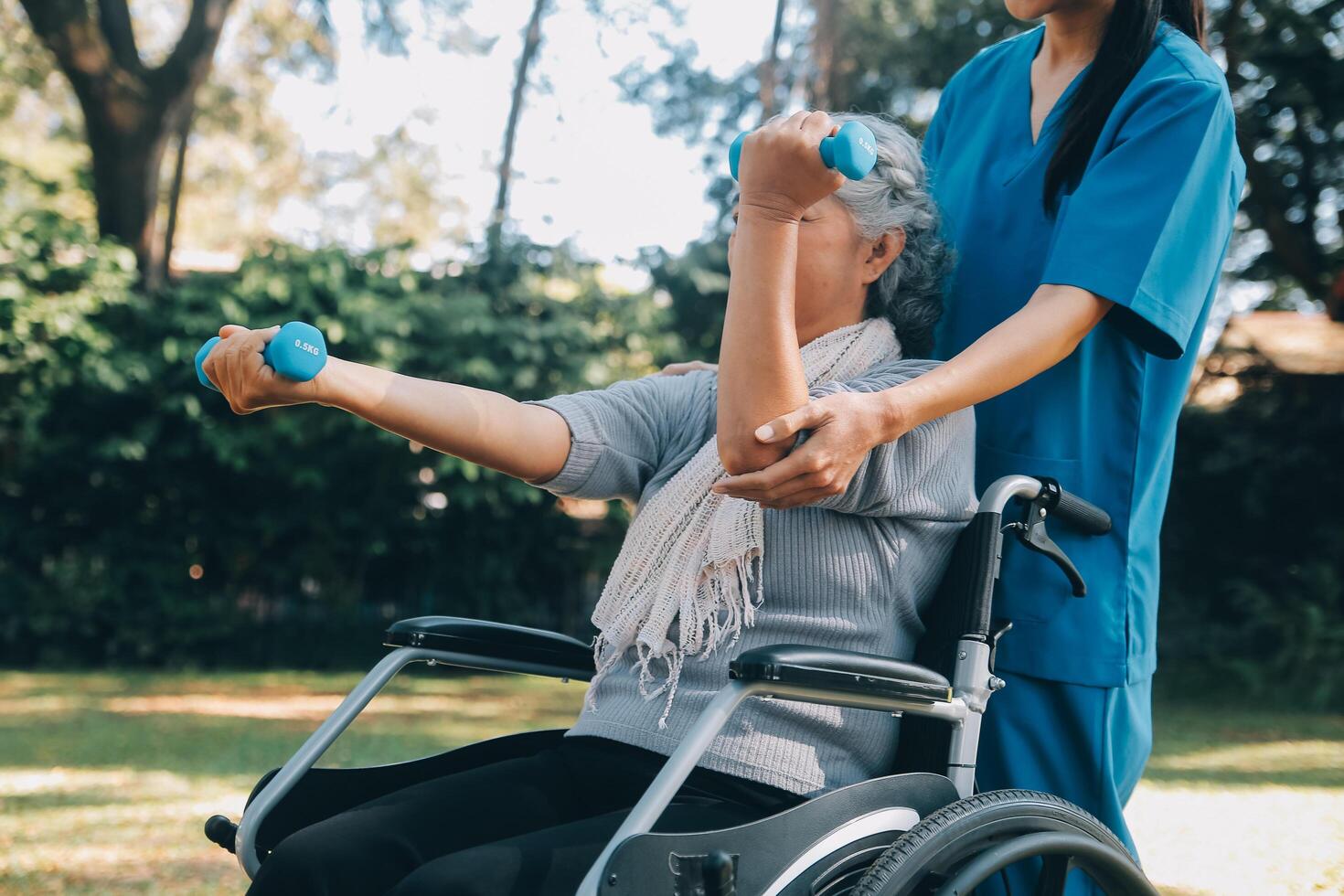 Young nurse or physiotherapist in scrubs helping a happy retired old woman do fitness exercises with light weight dumbbells at home. Concept of physiotherapy for seniors photo