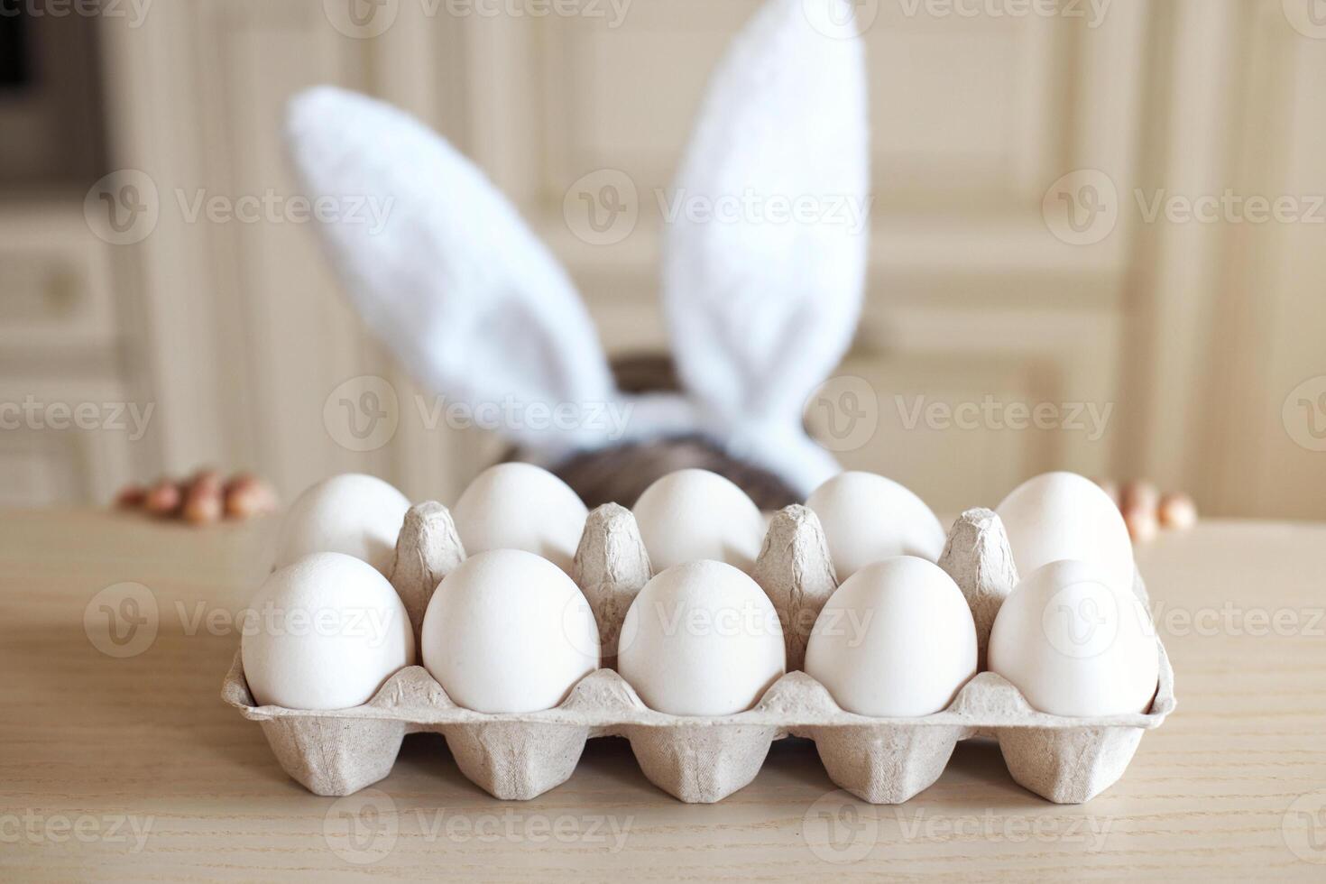 No face cute child with bunny ears is hiding under the table. White organic chicken eggs in a craft box stand on the table in the kitchen. Easter concept. photo