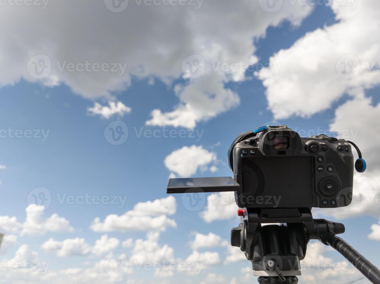 black professional digital camera with flip screen on a tripod pointed at blue sky with white clouds photo