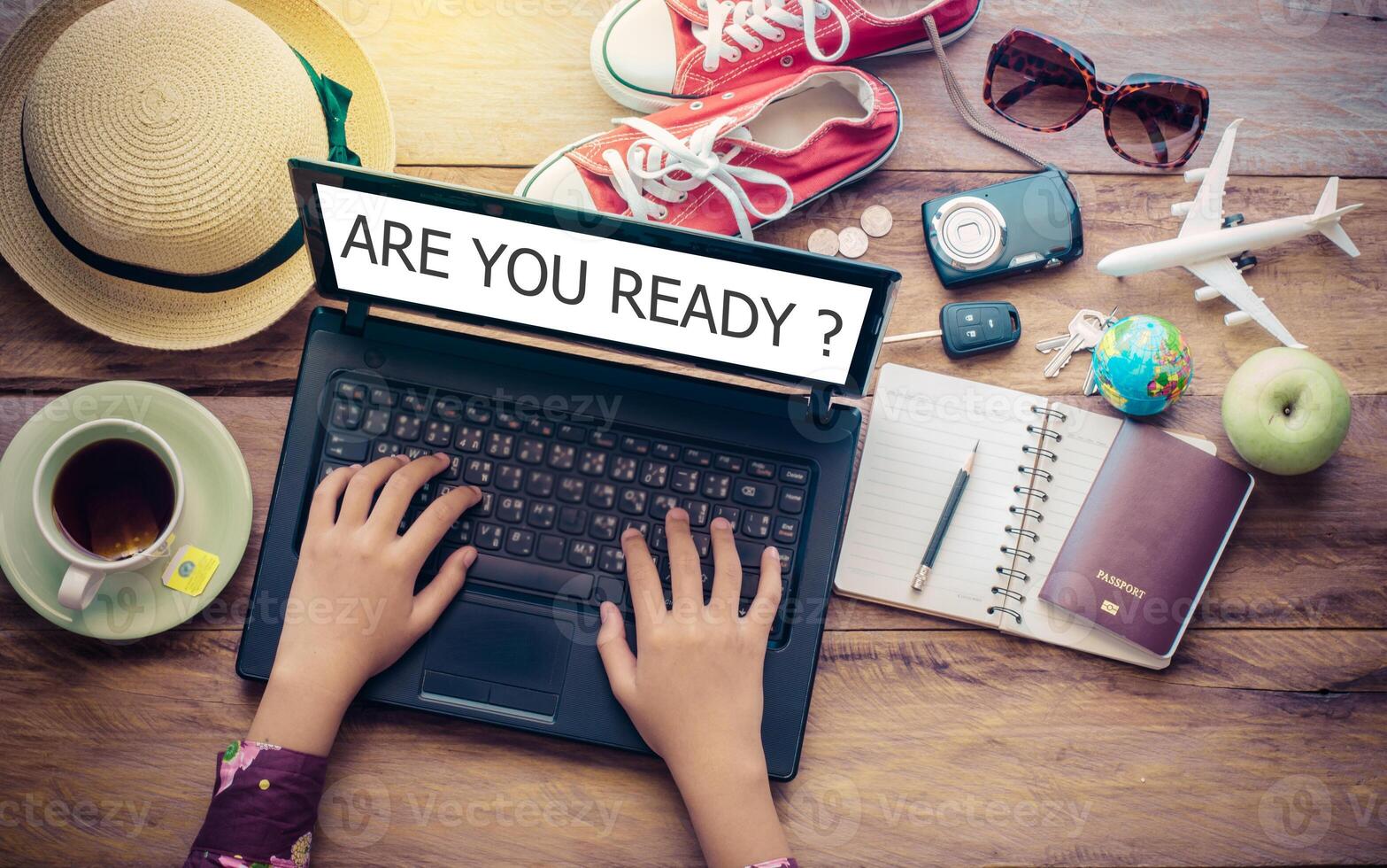 Hands on the keyboard of a notebook's screen displays, wo ARE YOU READY and travel accessories are placed on a wooden table. The concept plan tour photo