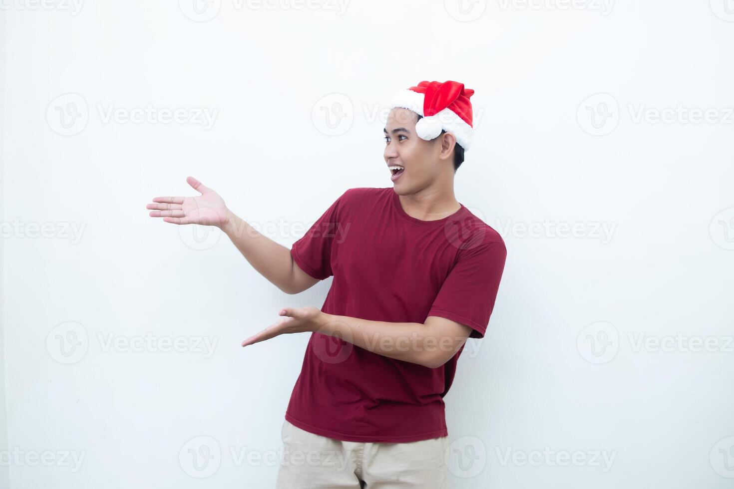 joven asiático hombre vistiendo un Papa Noel claus sombrero sonriente, conmoción y señalando a su lado aislado por blanco antecedentes para visual comunicación foto