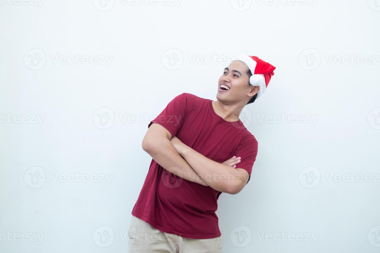 Young Asian man wearing a Santa Claus hat crossing his arms while smiling, shock and laughing with a white background for visual communication photo