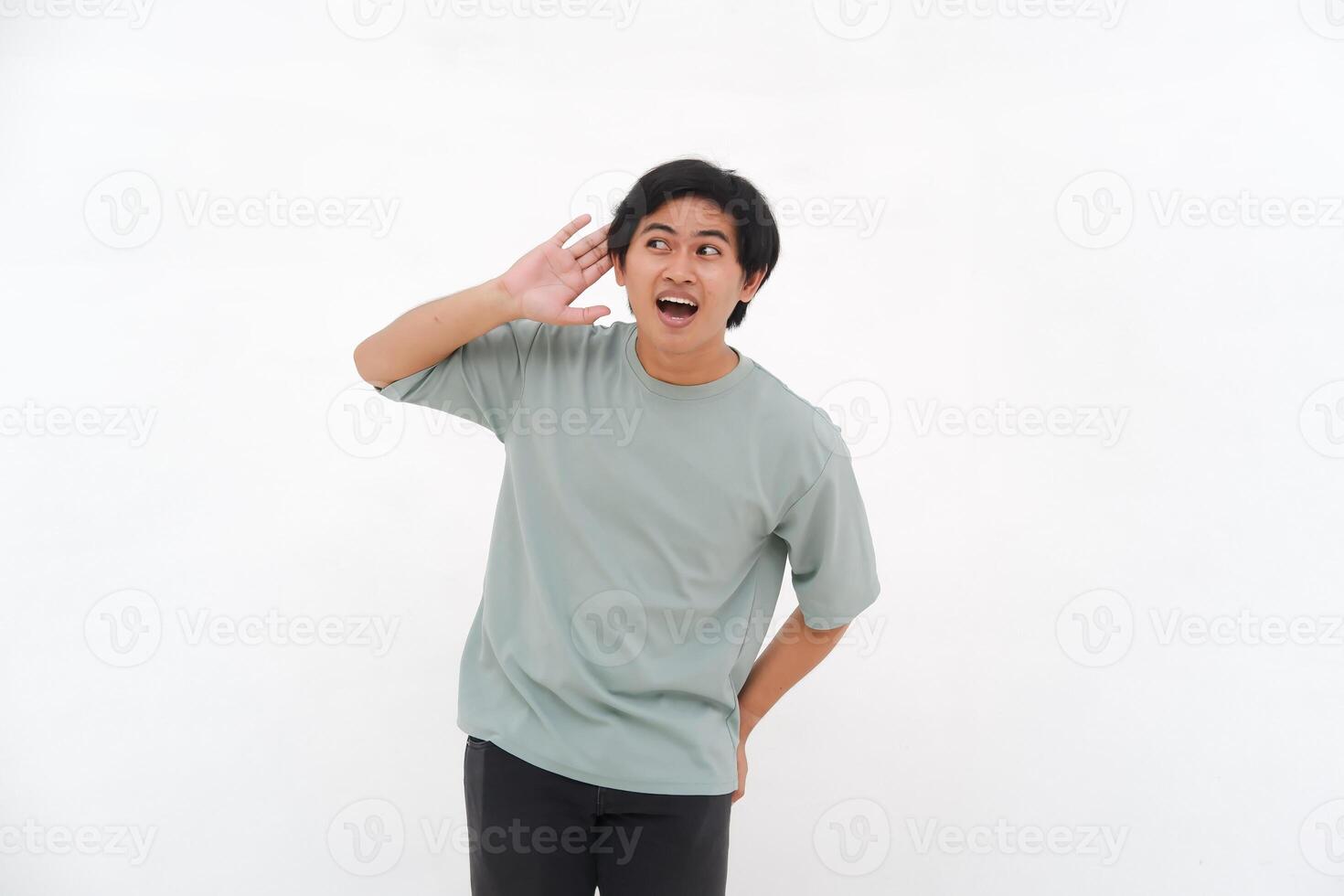 un joven asiático hombre, un empleado en un camiseta, es visto con entusiasmo escuchando con su mano a su oído en contra un blanco antecedentes. foto