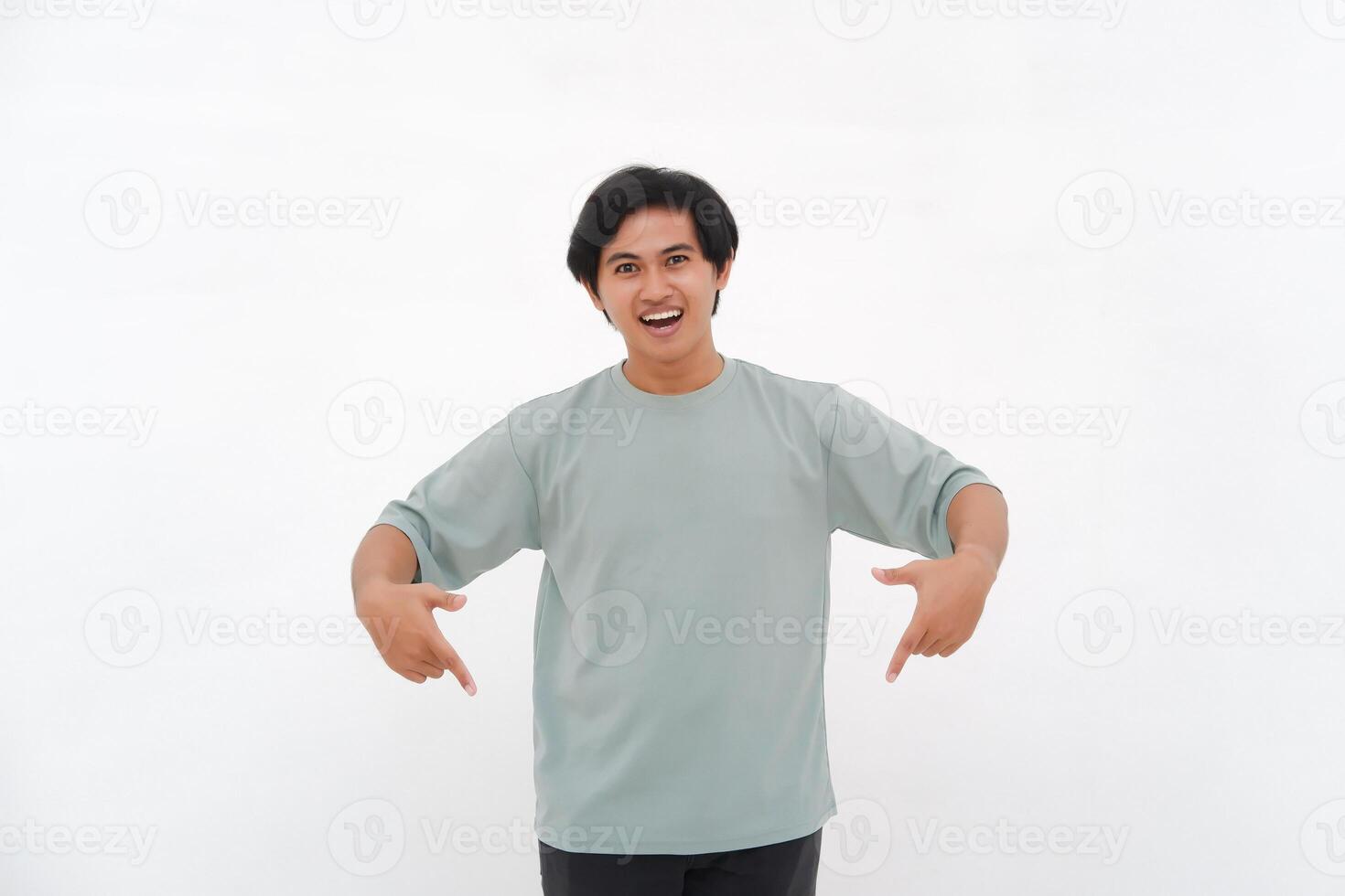 Excited Asian male employee wearing a T-shirt pointing at the copy space below him, isolated by a white background photo