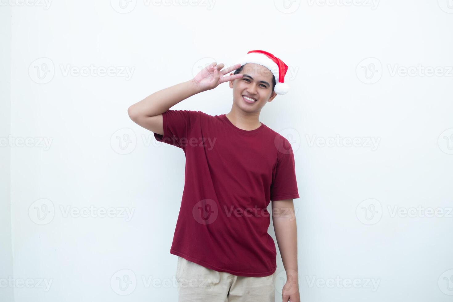 Young Asian man wearing a Santa Claus hat making a peace sign with 2 fingers isolated by a white background for visual communication photo