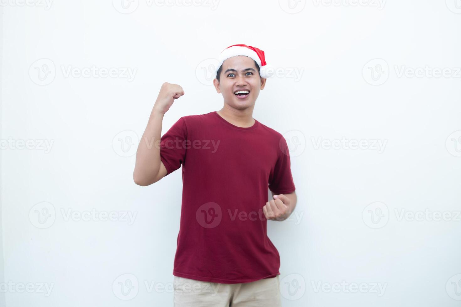 Young Asian man wearing a Santa Claus hat gripping both hands with enthusiasm and anger isolated by a white background for visual communication photo