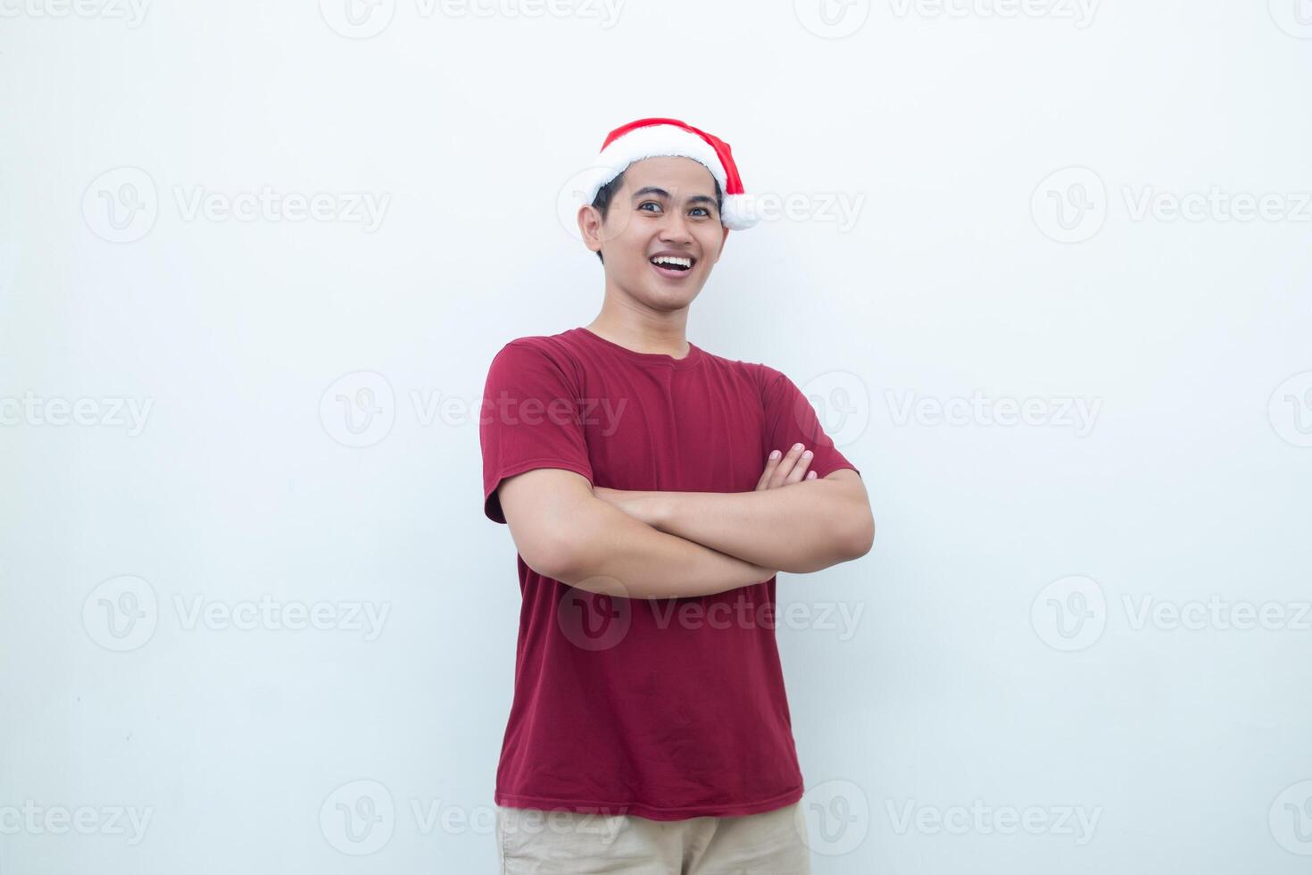 Young Asian man wearing a Santa Claus hat crossing his arms while smiling, shock and laughing with a white background for visual communication photo