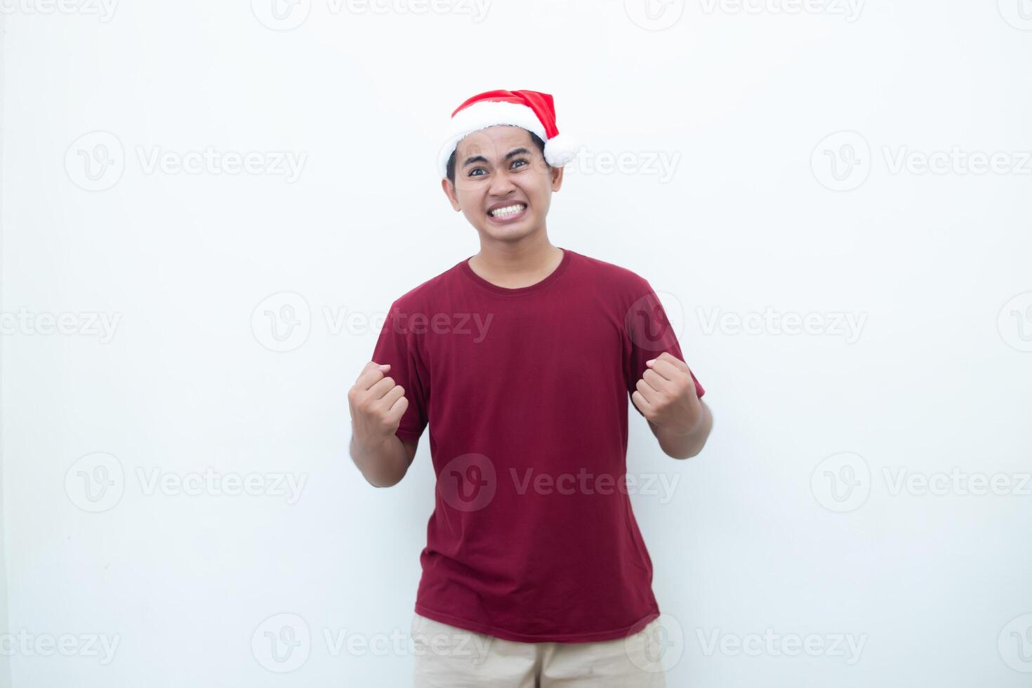 Young Asian man wearing a Santa Claus hat gripping both hands with enthusiasm and anger isolated by a white background for visual communication photo