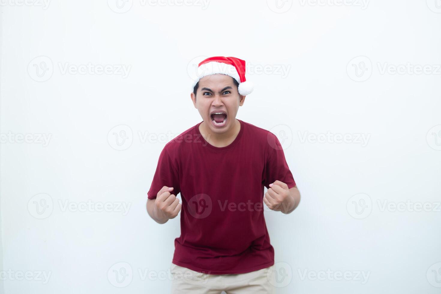 Young Asian man wearing a Santa Claus hat gripping both hands with enthusiasm and anger isolated by a white background for visual communication photo