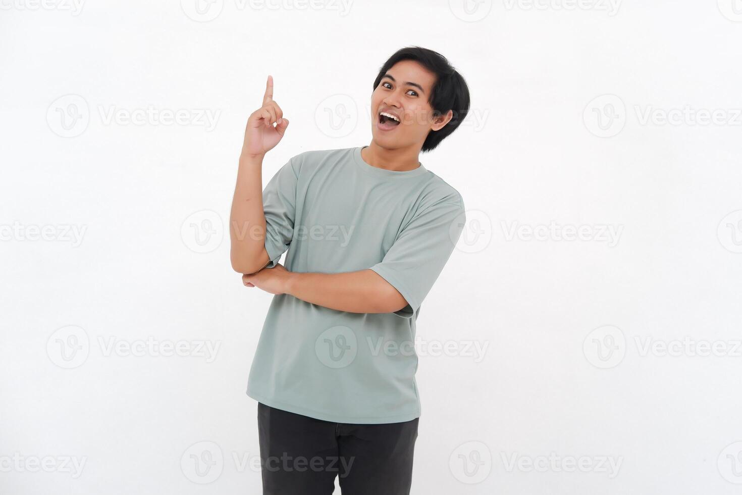 Excited Asian male employee wearing a tshirt pointing at the copy space above him, isolated by white background photo