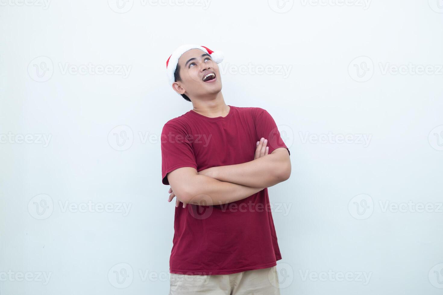 Young Asian man wearing a Santa Claus hat crossing his arms while smiling, shock and laughing with a white background for visual communication photo