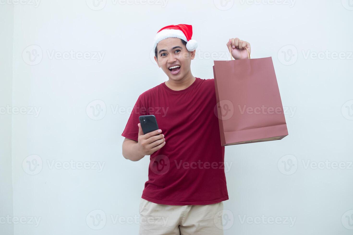 joven asiático hombre vistiendo un Papa Noel claus sombrero participación un teléfono inteligente y un compras bolso con expresiones de sonrisa, choque, y sorpresa, aislado en contra un blanco antecedentes para visual comunicación foto