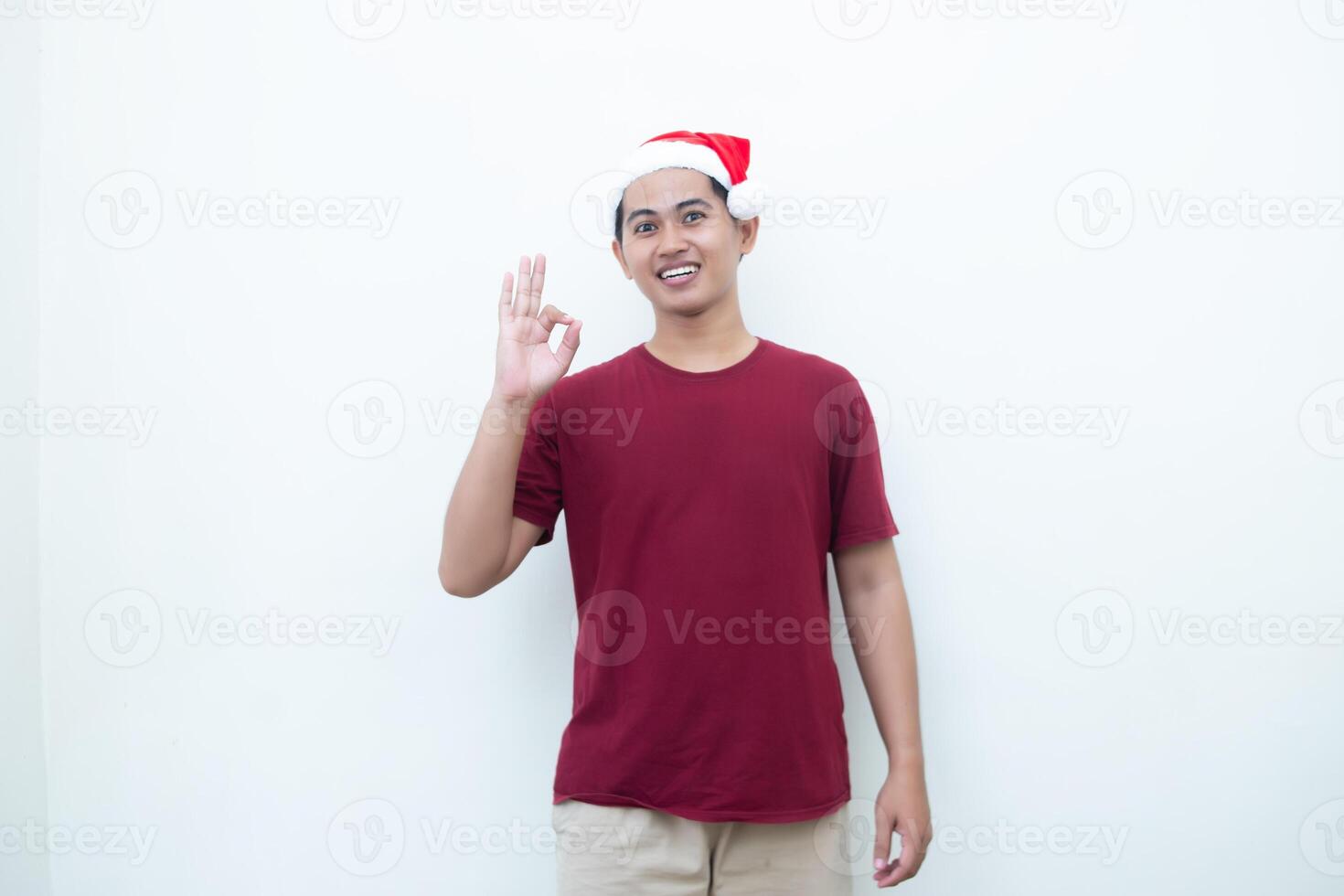 Young Asian man wearing a Santa Claus hat with a hand making the ok gesture isolated by a white background for visual communication photo