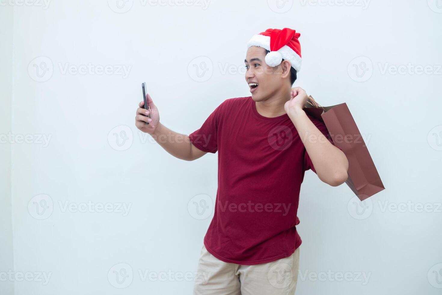 joven asiático hombre vistiendo un Papa Noel claus sombrero participación un teléfono inteligente y un compras bolso con expresiones de sonrisa, choque, y sorpresa, aislado en contra un blanco antecedentes para visual comunicación foto