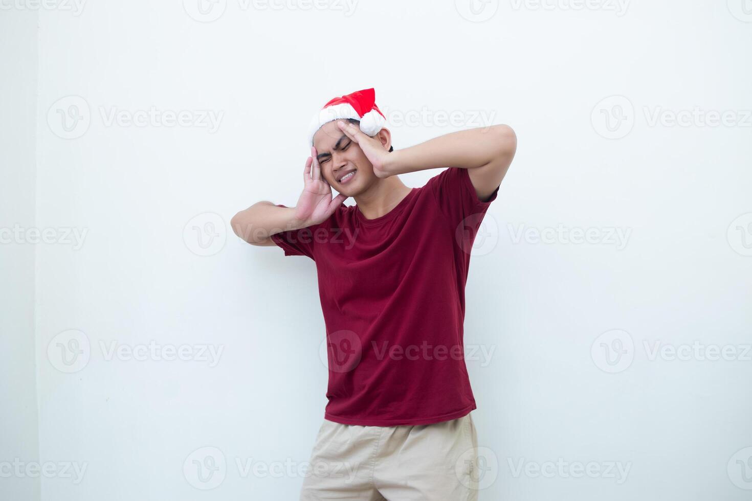 Young Asian man wearing a Santa Claus hat expressing a headache by holding his head and showing signs of pain isolated by a white background for visual communication photo