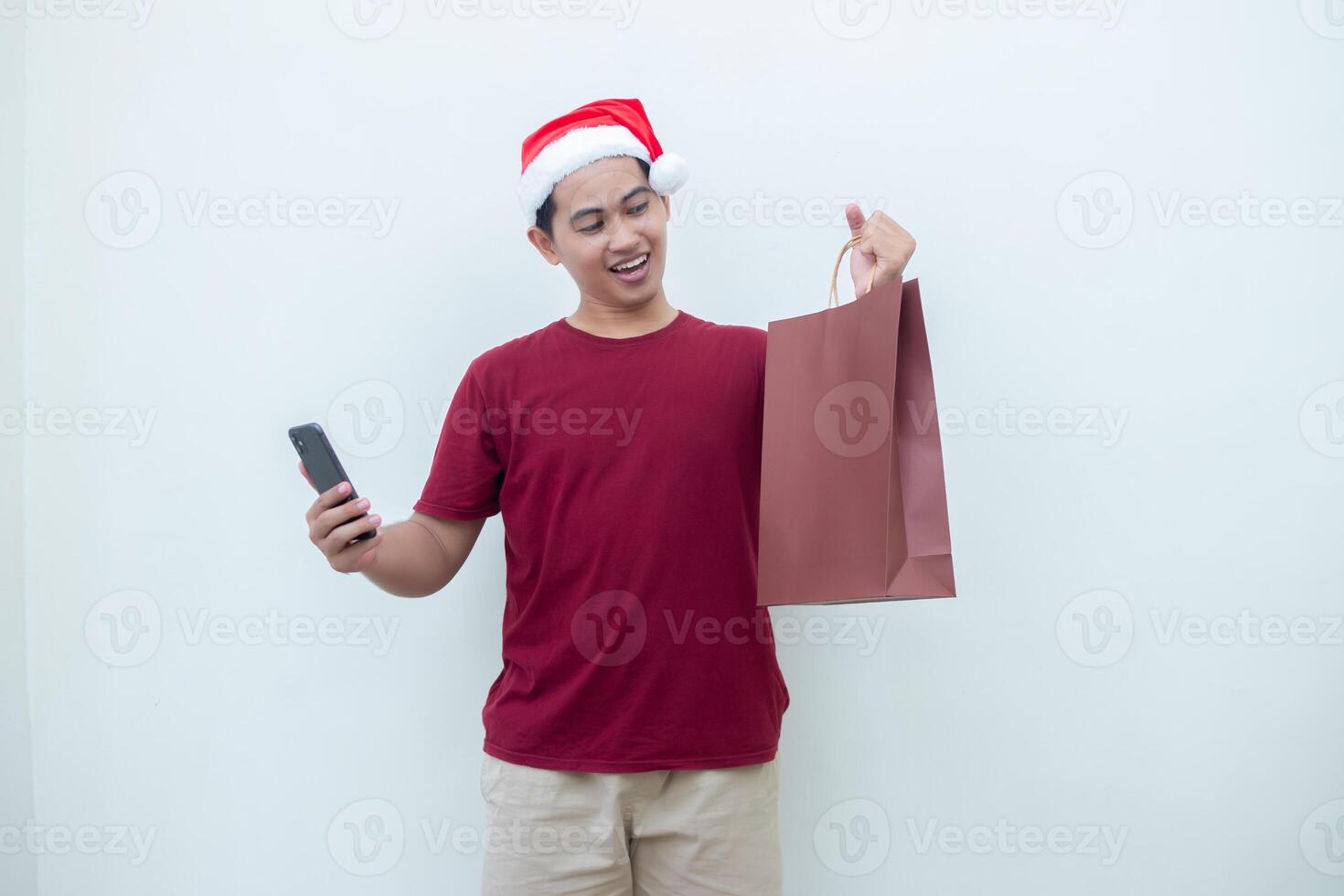 Young Asian man wearing a Santa Claus hat holding a smartphone and a shopping bag with expressions of smile, shock, and surprise, isolated against a white background for visual communication photo