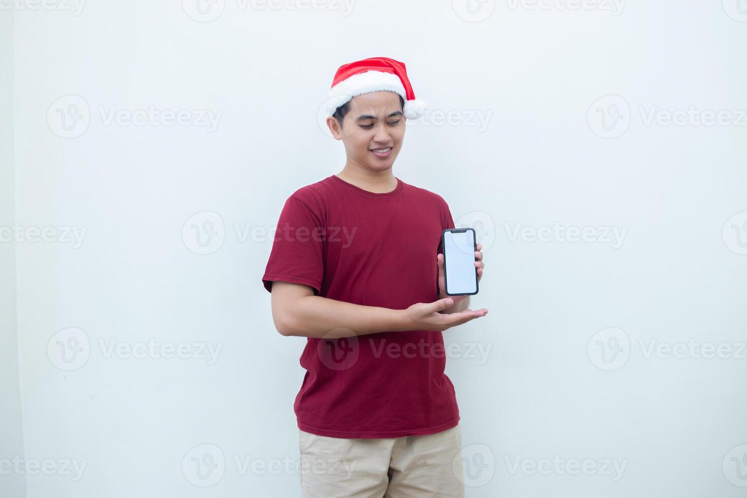 Young Asian man wearing a Santa Claus hat holding a smartphone, and pointing to the phone with a smile, look of shock, and surprise isolated against a white background for visual communication photo